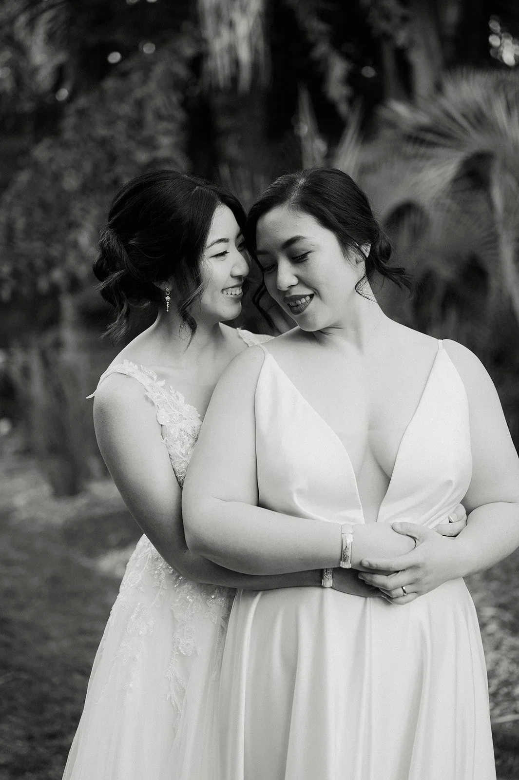 Two women in wedding dresses embracing and smiling in a natural outdoor setting with plants in the background.