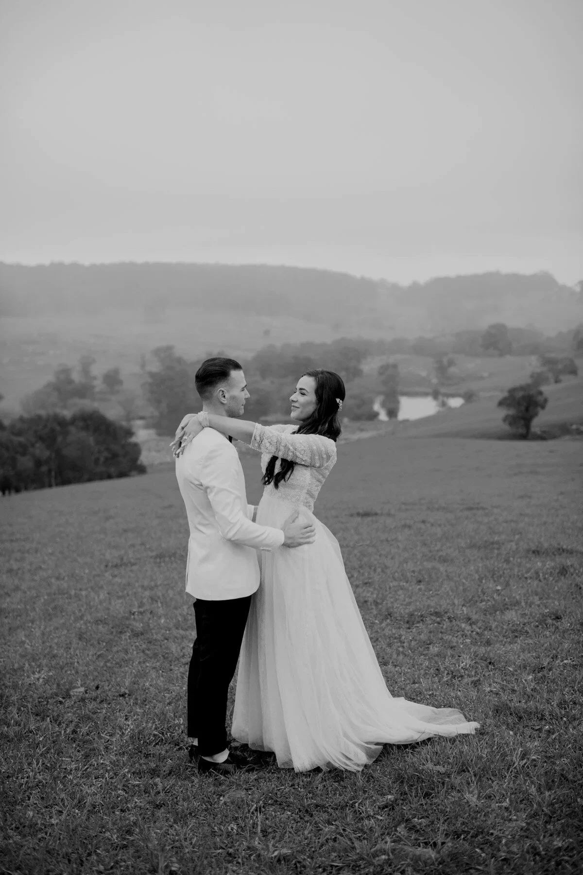 A black-and-white photo of a couple in wedding attire standing on a grassy field, embracing with rolling hills and sparse trees in the background.