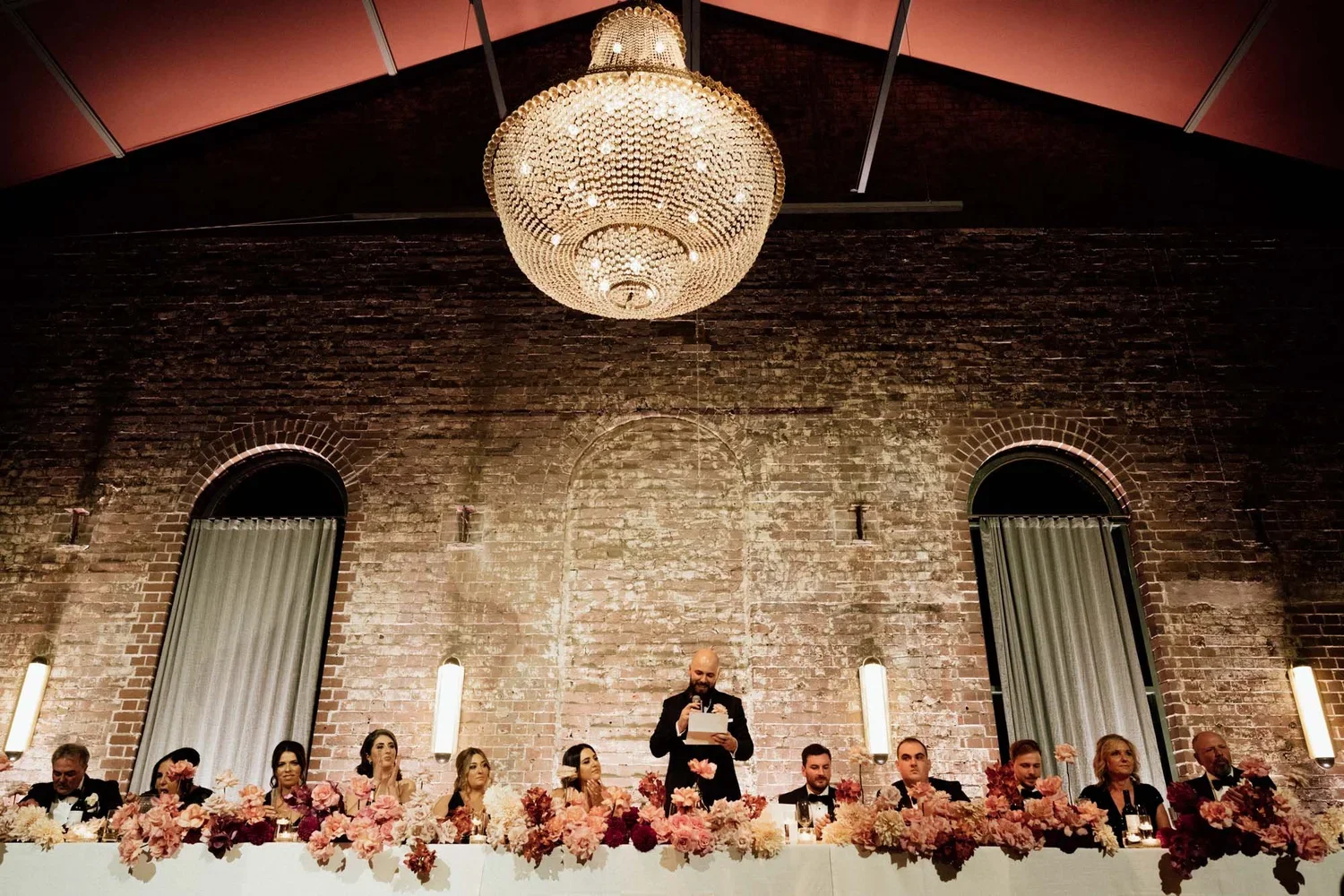 A wedding reception with a long head table decorated with pink and white flowers, behind which a man in a black tuxedo is giving a speech, flanked by bridesmaids and groomsmen dressed in formal attire, in a venue with exposed brick walls and modern l