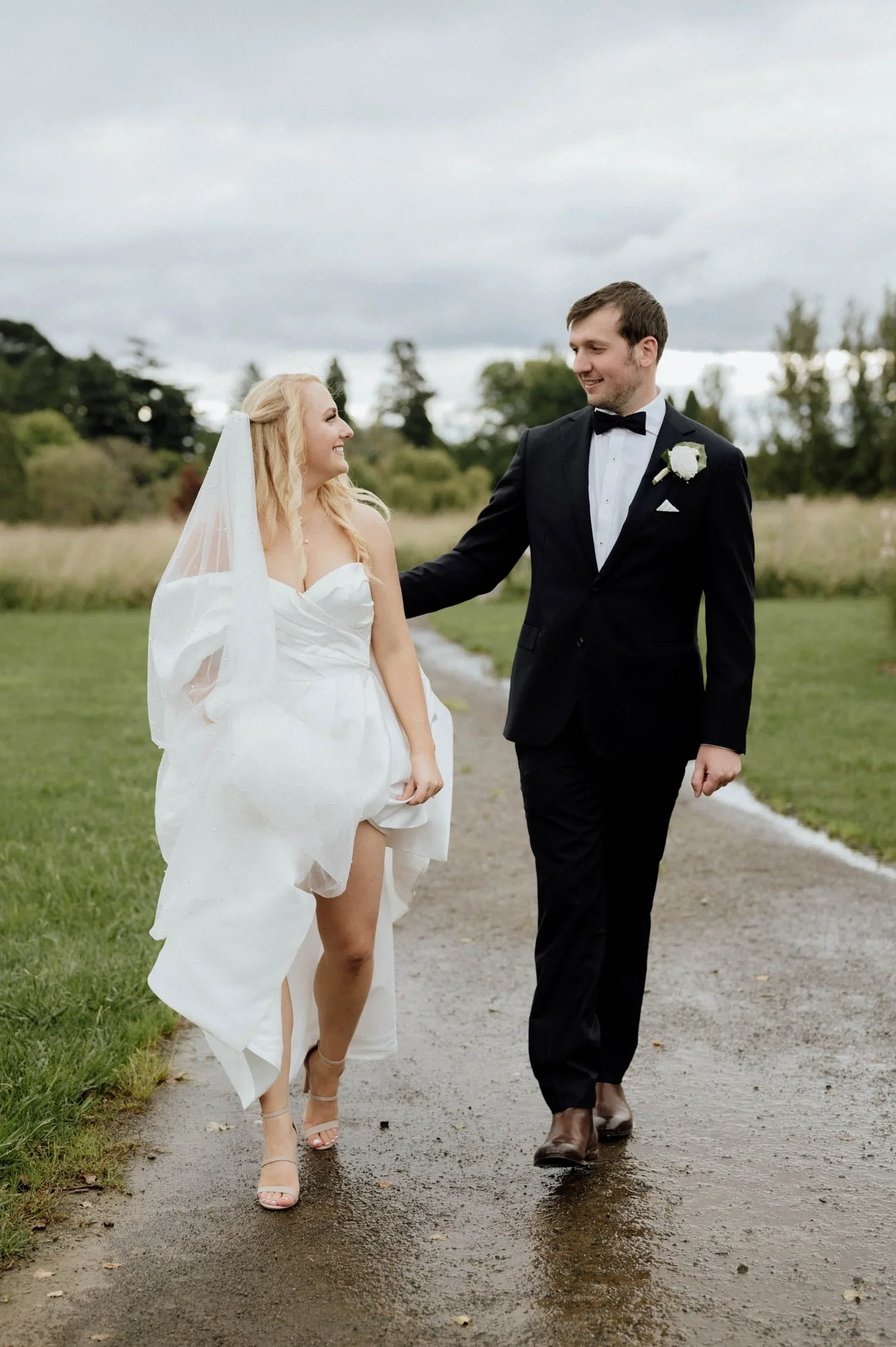 A bride in a white wedding dress with a veil and a groom in a black tuxedo walking together on a dirt path outdoors. The bride is smiling and looking at the groom, while the groom is smiling and looking at the bride. The sky is cloudy and there's gre