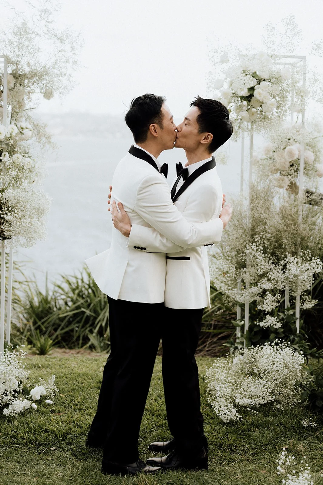 Two grooms at their same sex wedding in tuxedos embracing and kissing at a wedding ceremony outdoors, with white floral arrangements and greenery surrounding them.