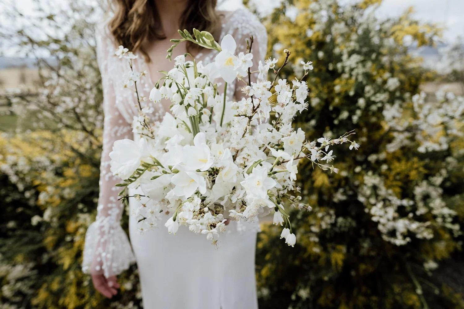 A woman in a white dress holds a bouquet of white flowers, with yellow and green accents, in front of a bush with white flowers and yellow-green leaves.