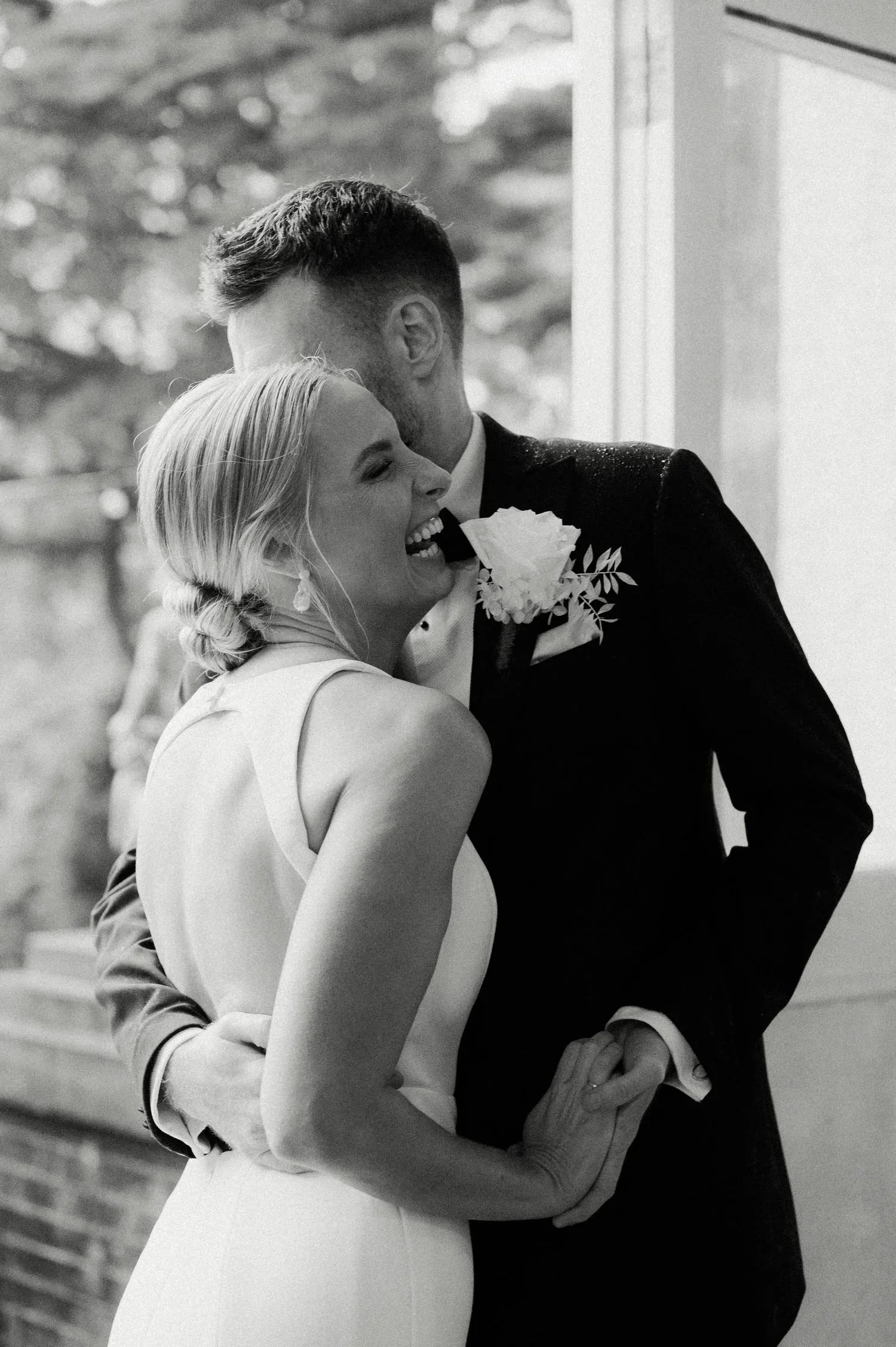 A couple is dancing closely, with the woman smiling and the man with his eyes closed, wearing formal clothing, against a blurred background.