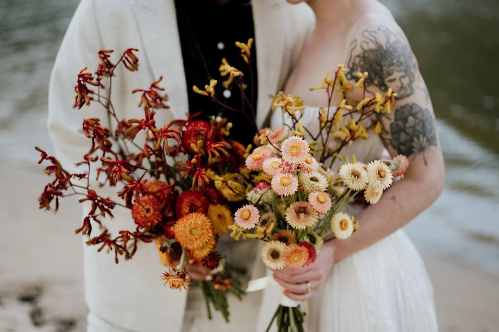 Two brides holding their bouquet, lesbian wedding couple in a white dress holding a bouquet of orange, pink, and yellow flowers near the water.