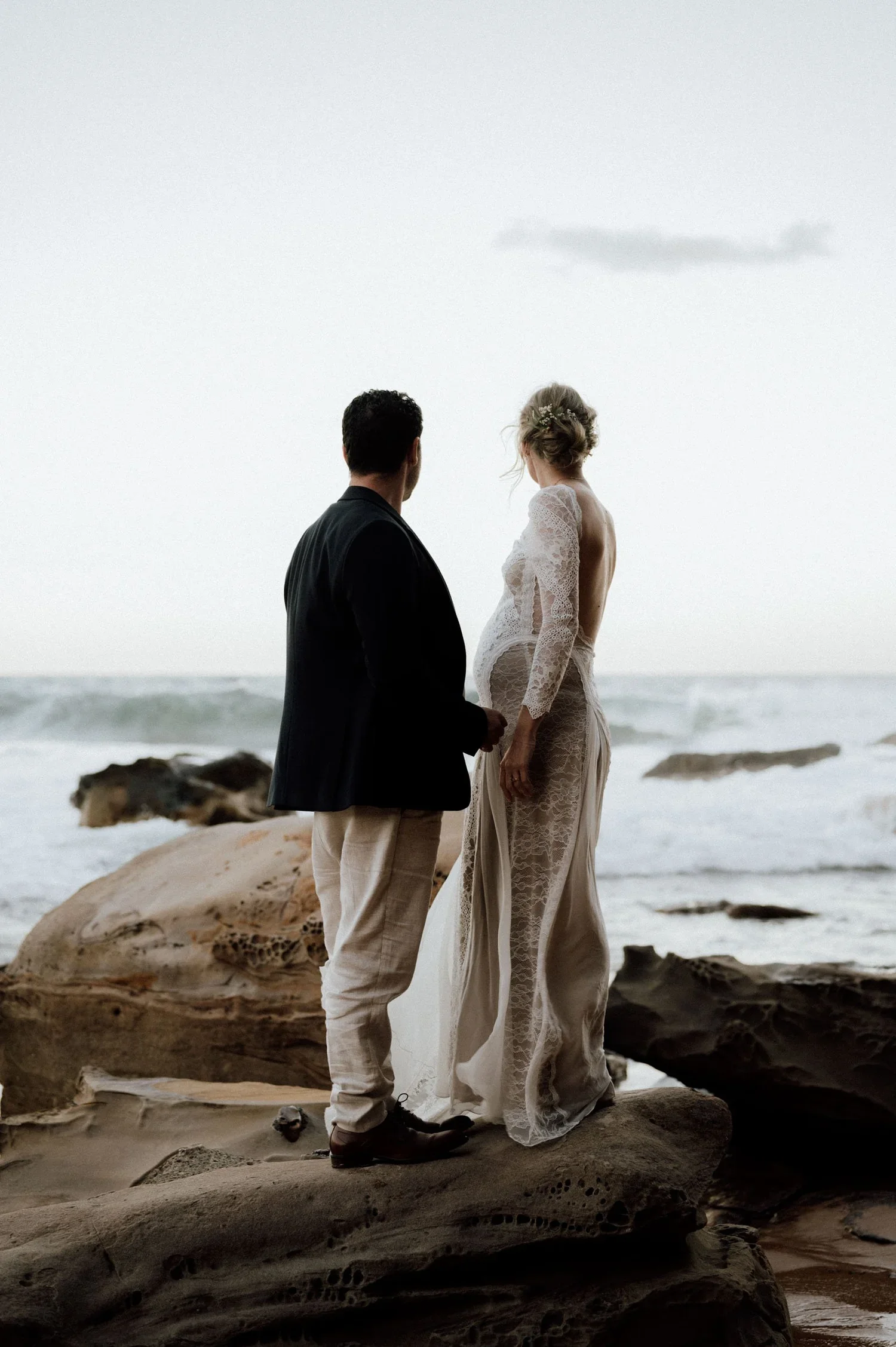 A couple dressed in wedding attire standing on rocks on the beach at sunset, facing each other with the ocean and sky in the background.