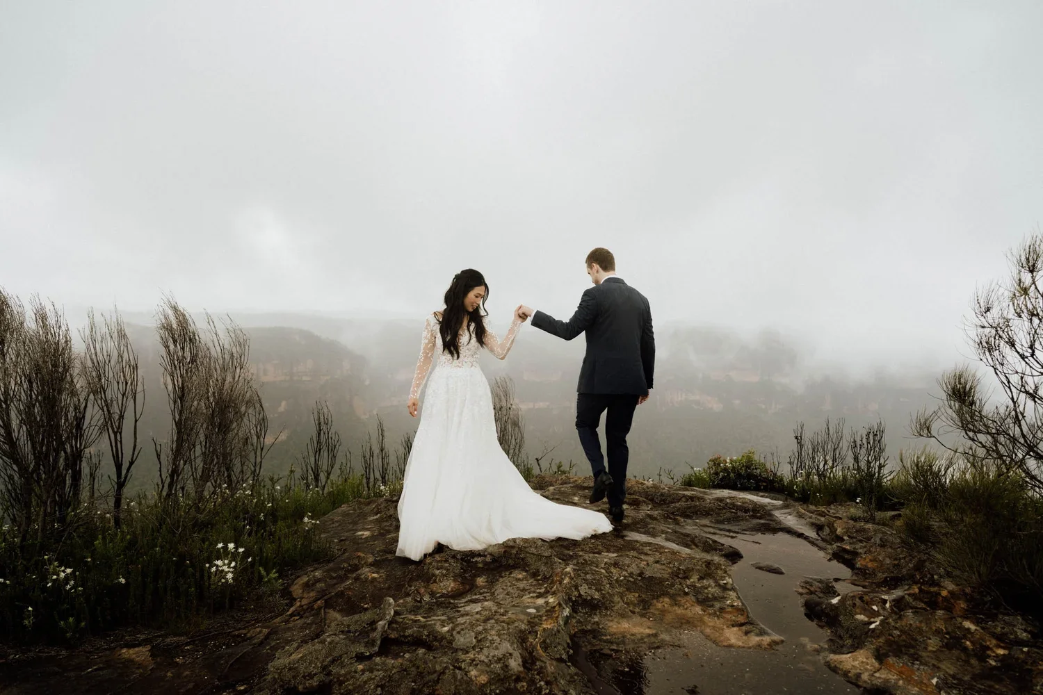 A bride and groom holding hands on a rocky hilltop with a misty, foggy landscape in the background, fog obscuring distant hills.