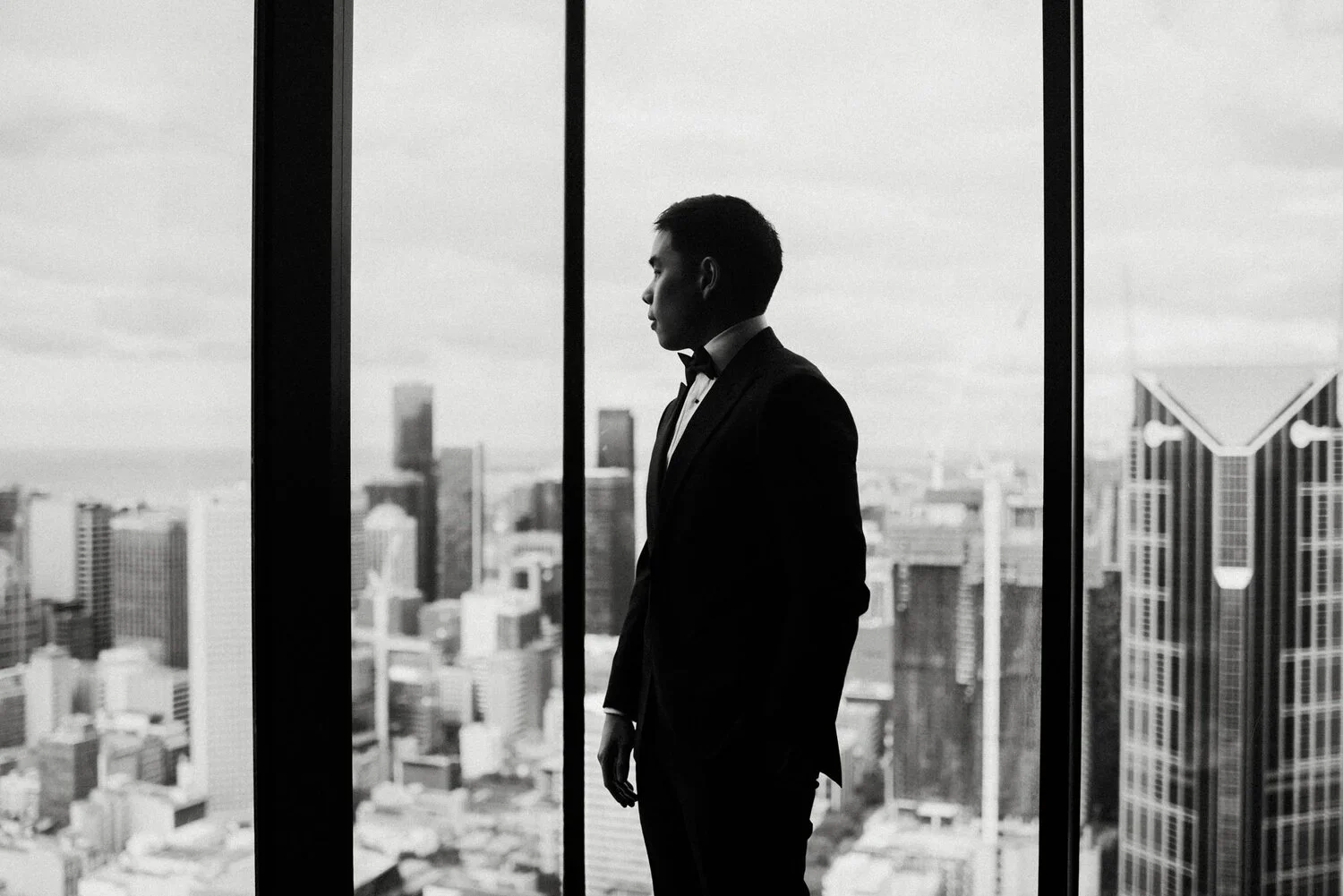 Silhouette of a man in a tuxedo standing in front of large windows overlooking a city skyline in black and white.