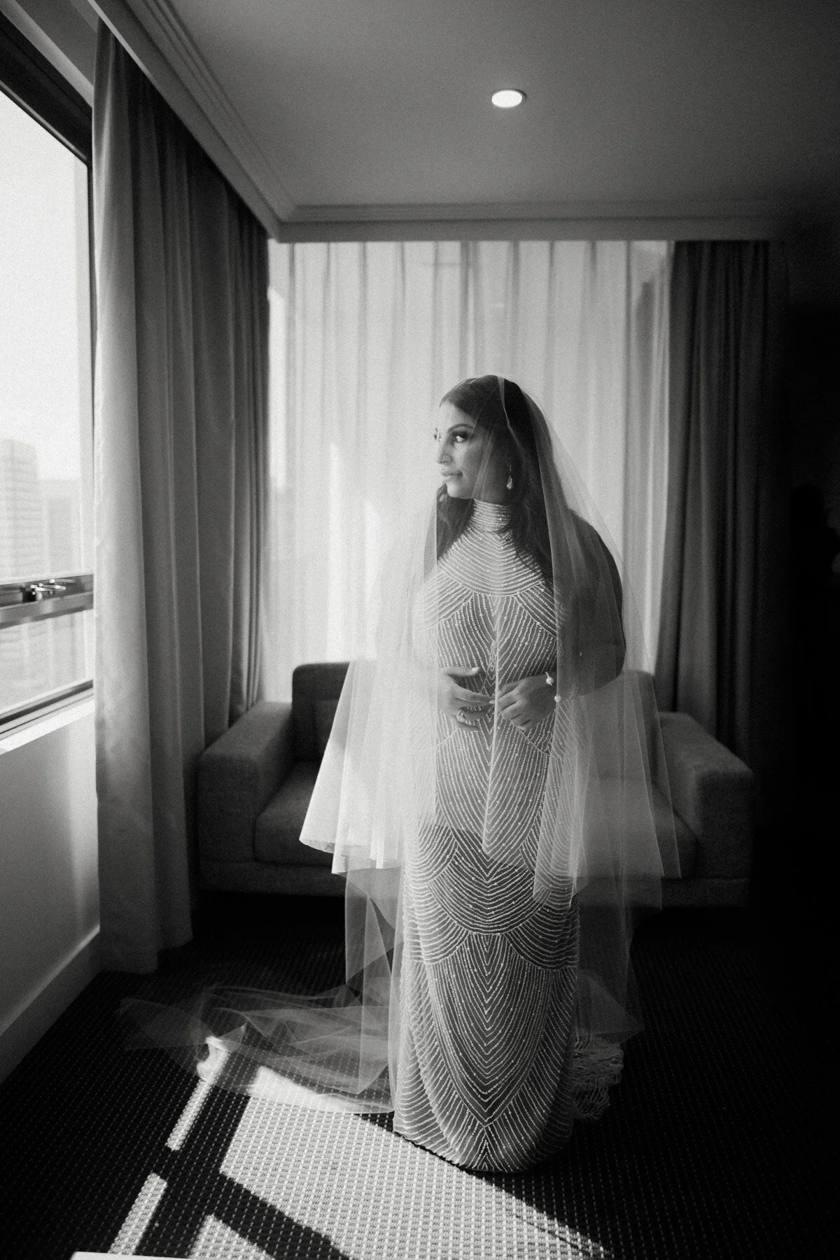 A bride in a patterned wedding dress with a veil, standing indoors near a window with curtains, holding a glass, with a sofa and window view in the background.