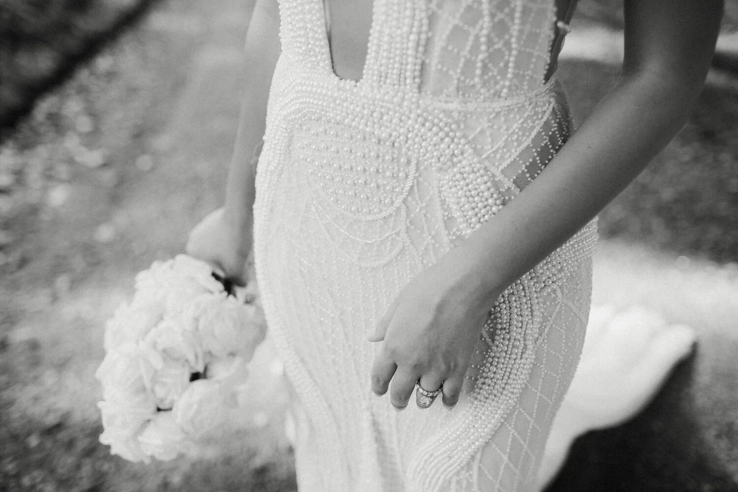 A close-up of a woman in a pearl-adorned wedding dress holding a bouquet of flowers, showing her jewelry on her left hand.
