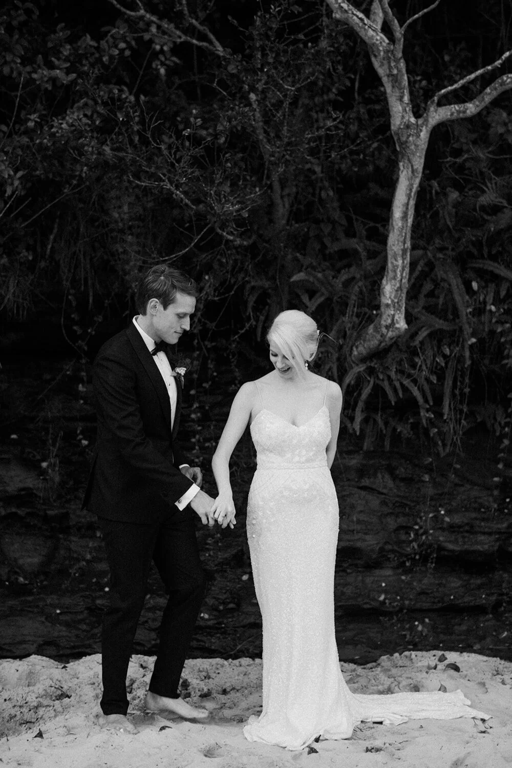 A black and white photo of a couple dressed in wedding attire holding hands and smiling on a sandy beach with trees and foliage in the background.