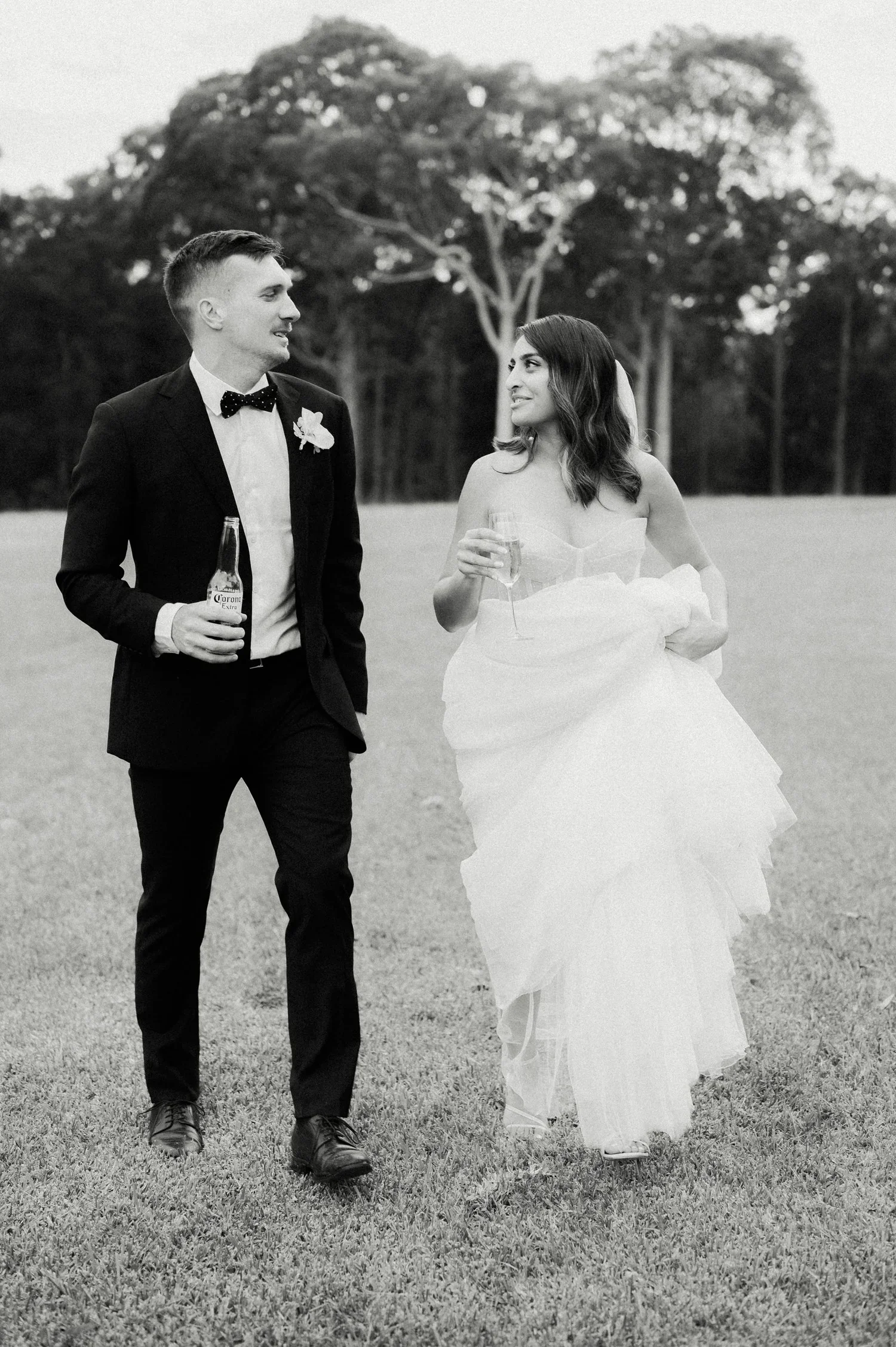 Black and white photo of a newlywed couple walking across a grassy field, holding drinks and smiling at each other, with trees in the background.