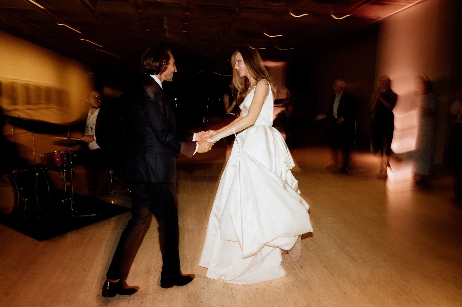 A bride and groom dancing together at their wedding reception, holding hands and smiling, with guests in the background.