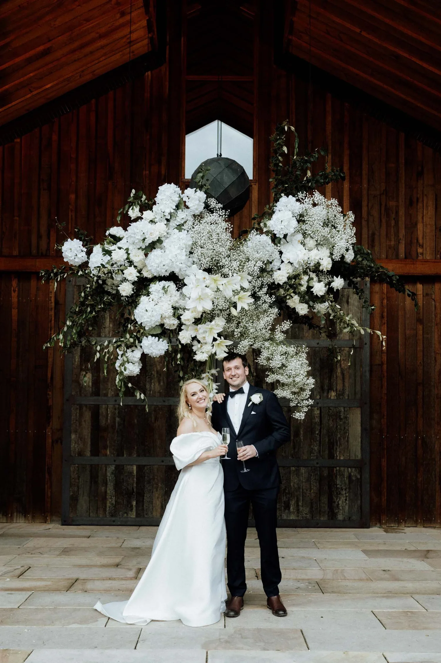 A newlywed couple standing in front of a large floral arrangement inside a barn. The bride is wearing a white wedding gown and holding a glass of champagne, and the groom is dressed in a black tuxedo with a bow tie, also holding a glass of champagne,