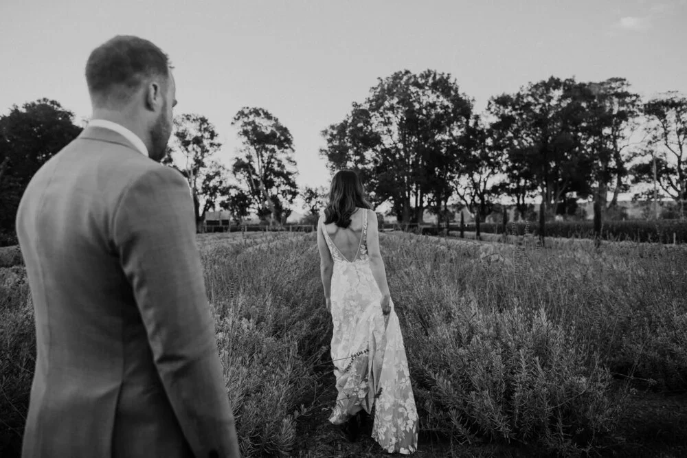 A man and woman in formal attire standing outdoors in a field with trees in the background during sunset, with the woman walking ahead.