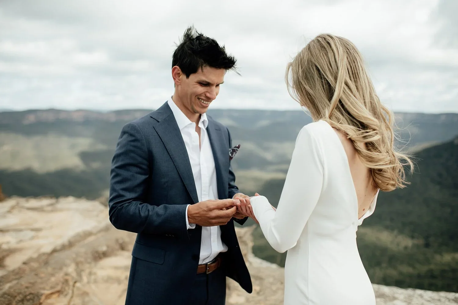 A man in a suit exchanging rings with a woman in a white dress during a wedding ceremony outdoors with mountain views.
