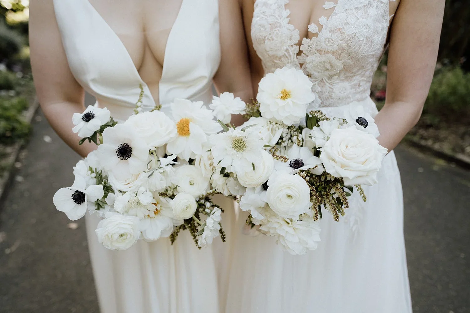 Two women wearing wedding dresses holding bouquets of white flowers, including roses and anemones, outdoors.