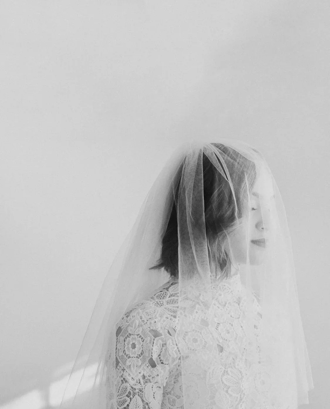 A woman in a wedding dress with lace details, wearing a veil, standing sideways with her eyes closed against a plain white wall.