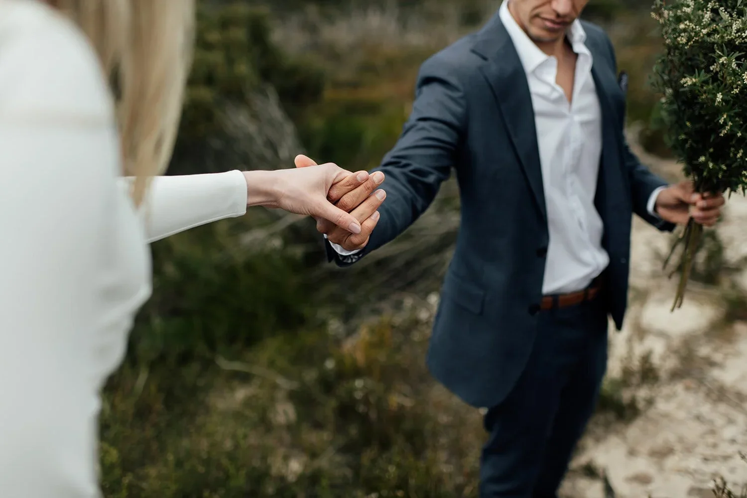 A man in a suit holding a bouquet of flowers, reaching out his hand to a woman in a white dress amid an outdoor setting.