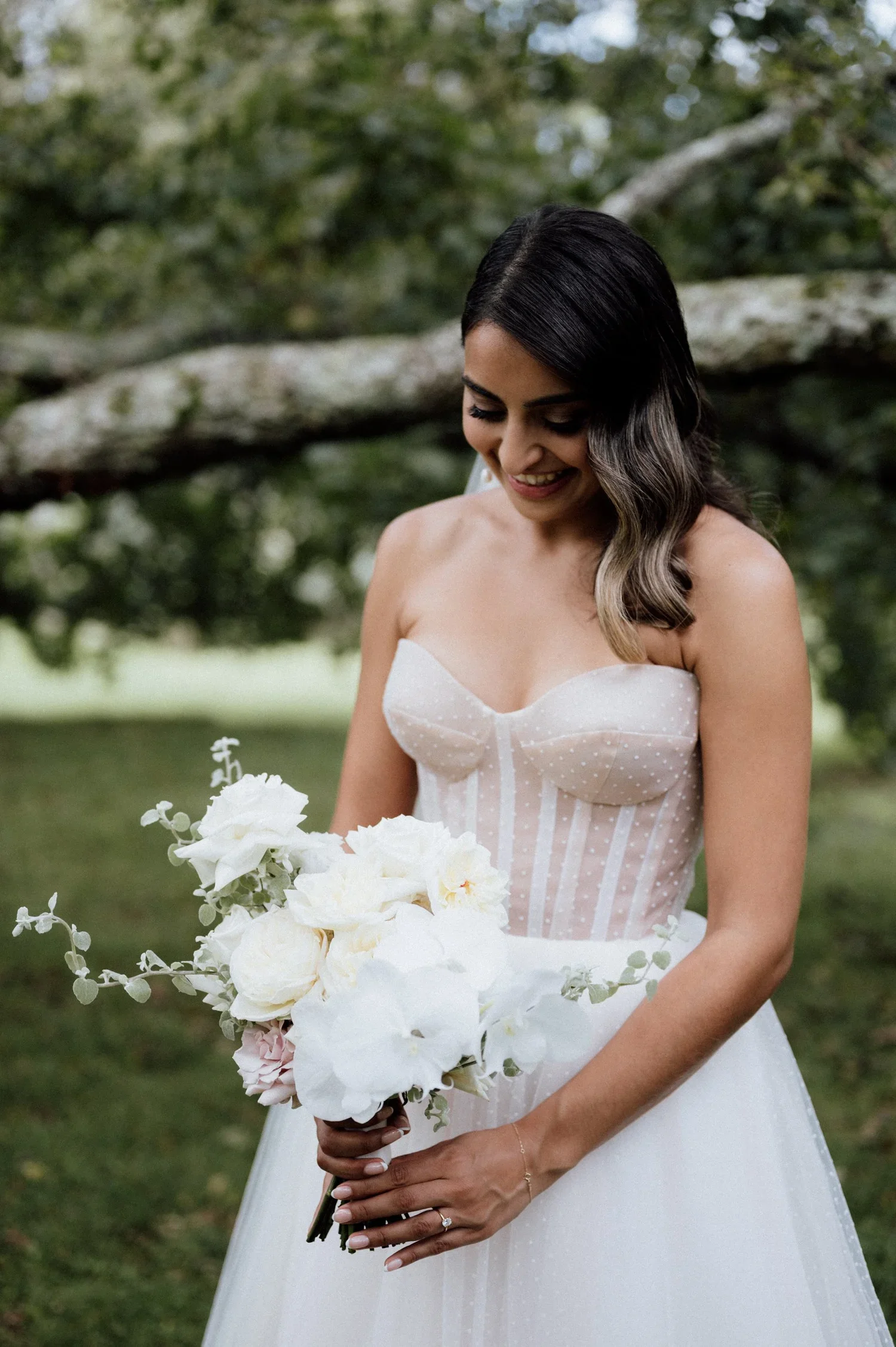 A woman in a white dress holding a bouquet of white flowers outdoors with green trees in the background.