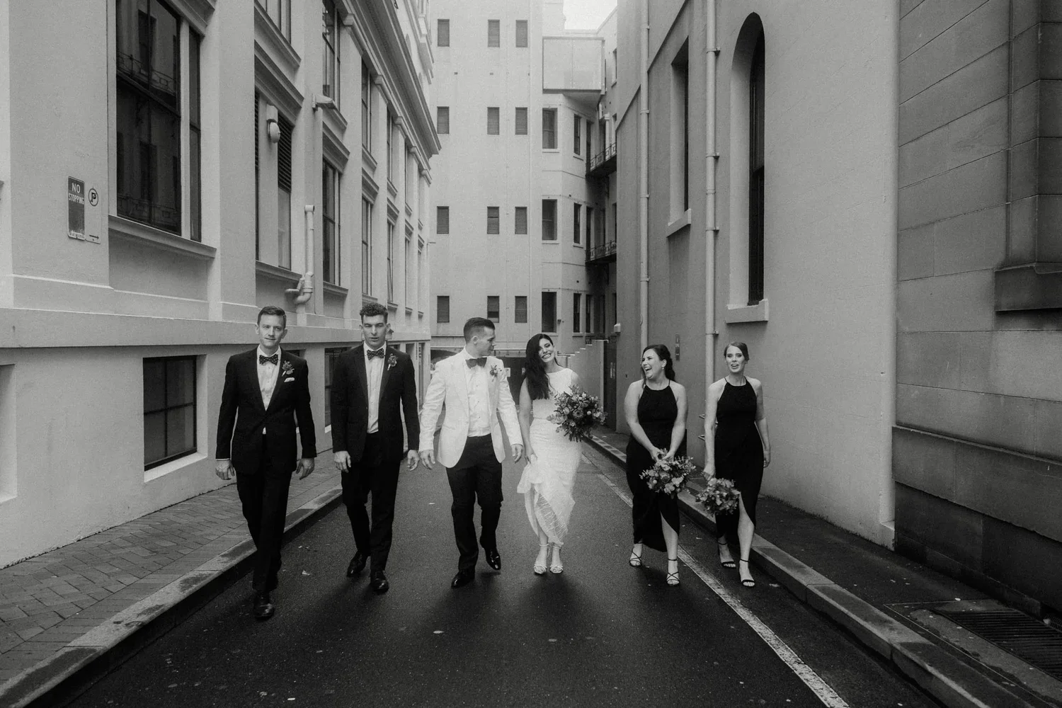 A wedding party walking down a city alleyway, including a bride in a white dress holding a bouquet, a groom in a light suit, and four bridesmaids and groomsmen dressed in formal attire.
