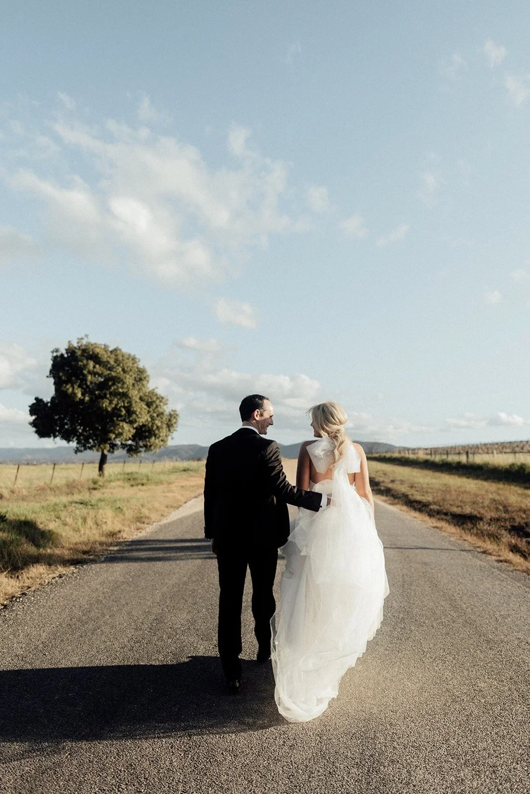 A couple in wedding attire walking hand in hand down a rural road on a sunny day, with open fields, a large tree, and a blue sky with scattered clouds in the background - Mudgee wedding venue and photography.