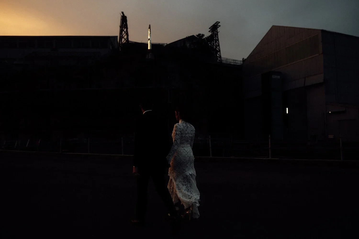 A man and woman dressed in formal attire walking outdoors during dusk, with industrial structures and a building in the background.