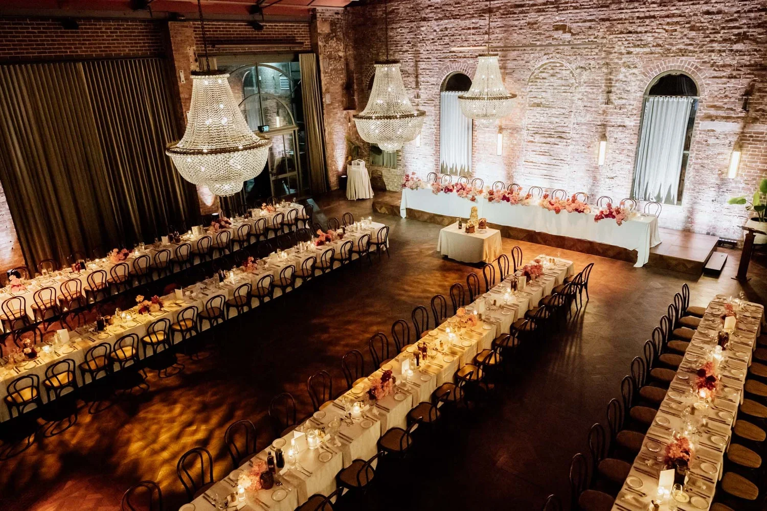 Elegant banquet hall decorated for a wedding reception with long tables set with white tablecloths, pink and purple floral centerpieces, candles, and place settings. Large crystal chandeliers hang from exposed brick walls, with tall windows partially