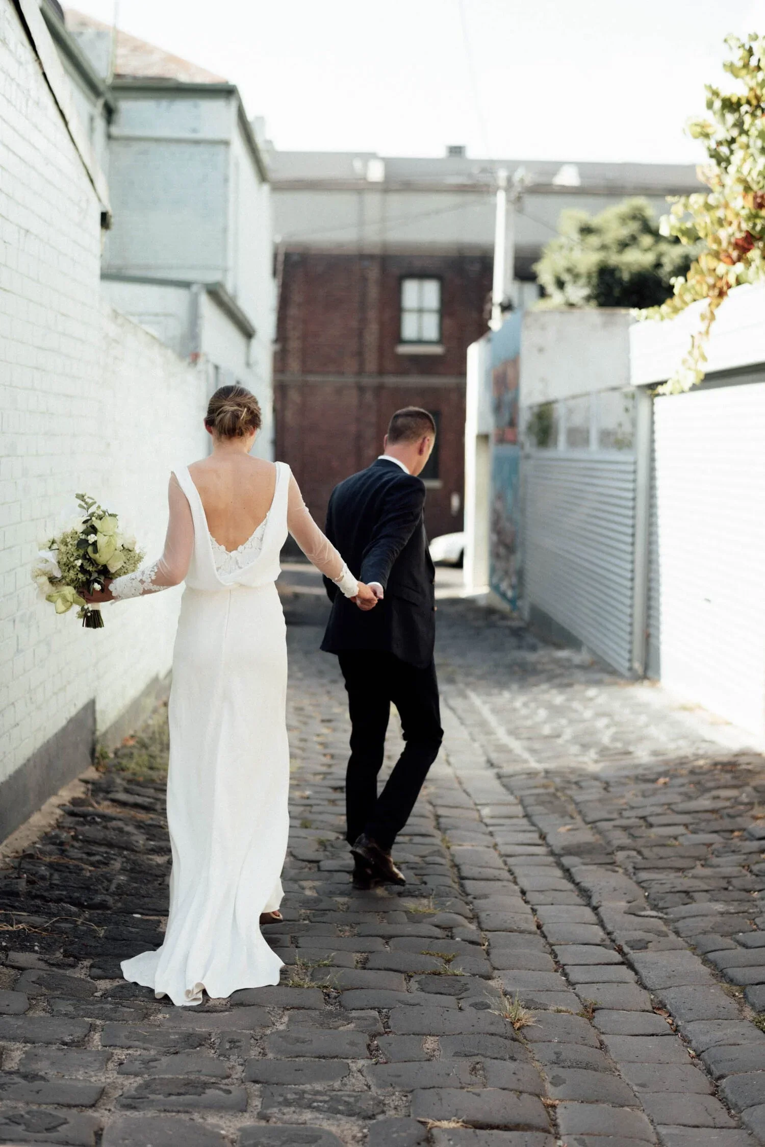 A bride and groom holding hands and walking along a cobblestone alleyway, with the bride wearing a white wedding gown holding a bouquet and the groom in a black suit.