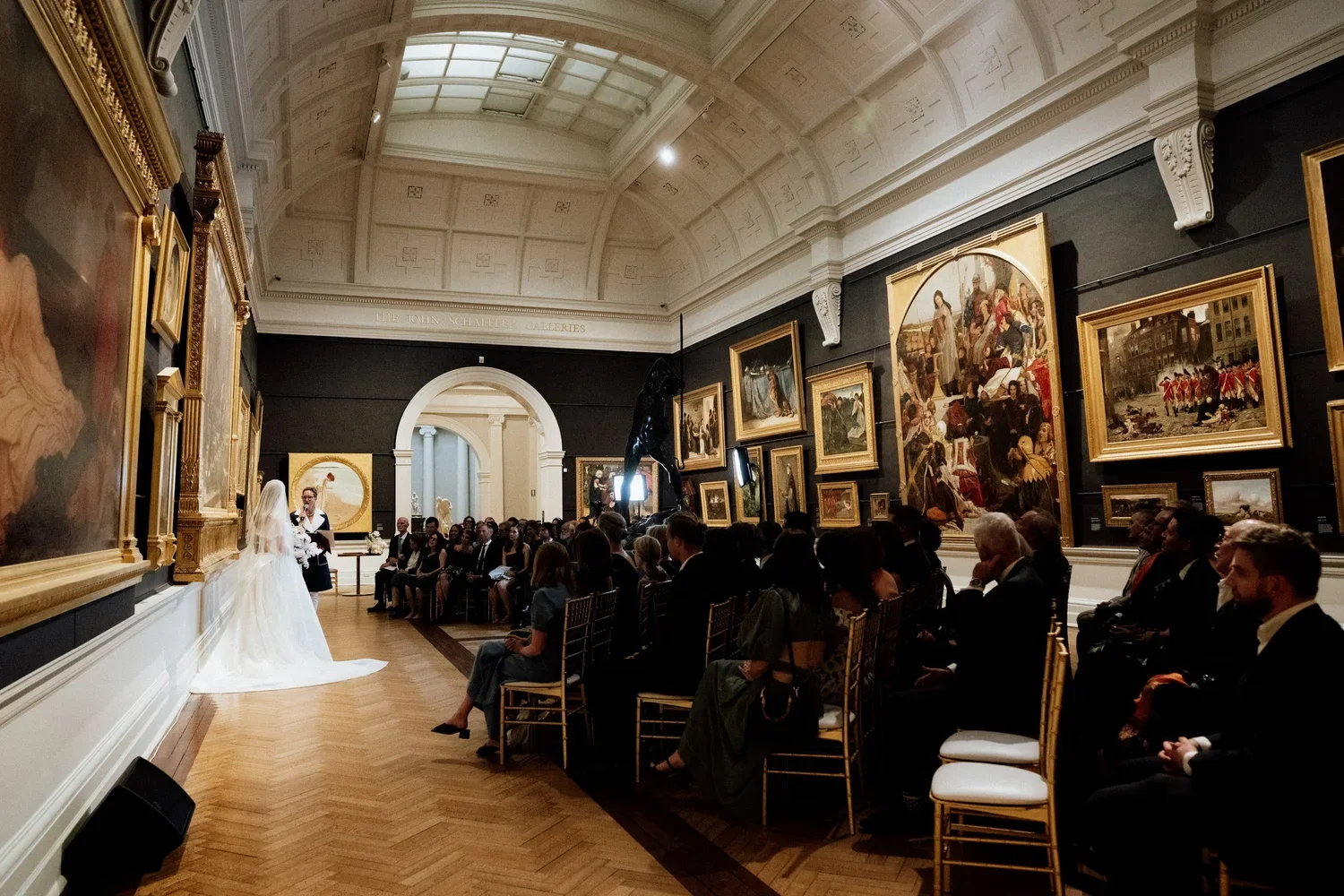 A wedding ceremony taking place in art gallery with paintings on the walls and a high, curved ceiling. The bride in a white gown and veil is standing with a woman in black and white, possibly the officiant. Guests are seated on gold chairs, watching 
