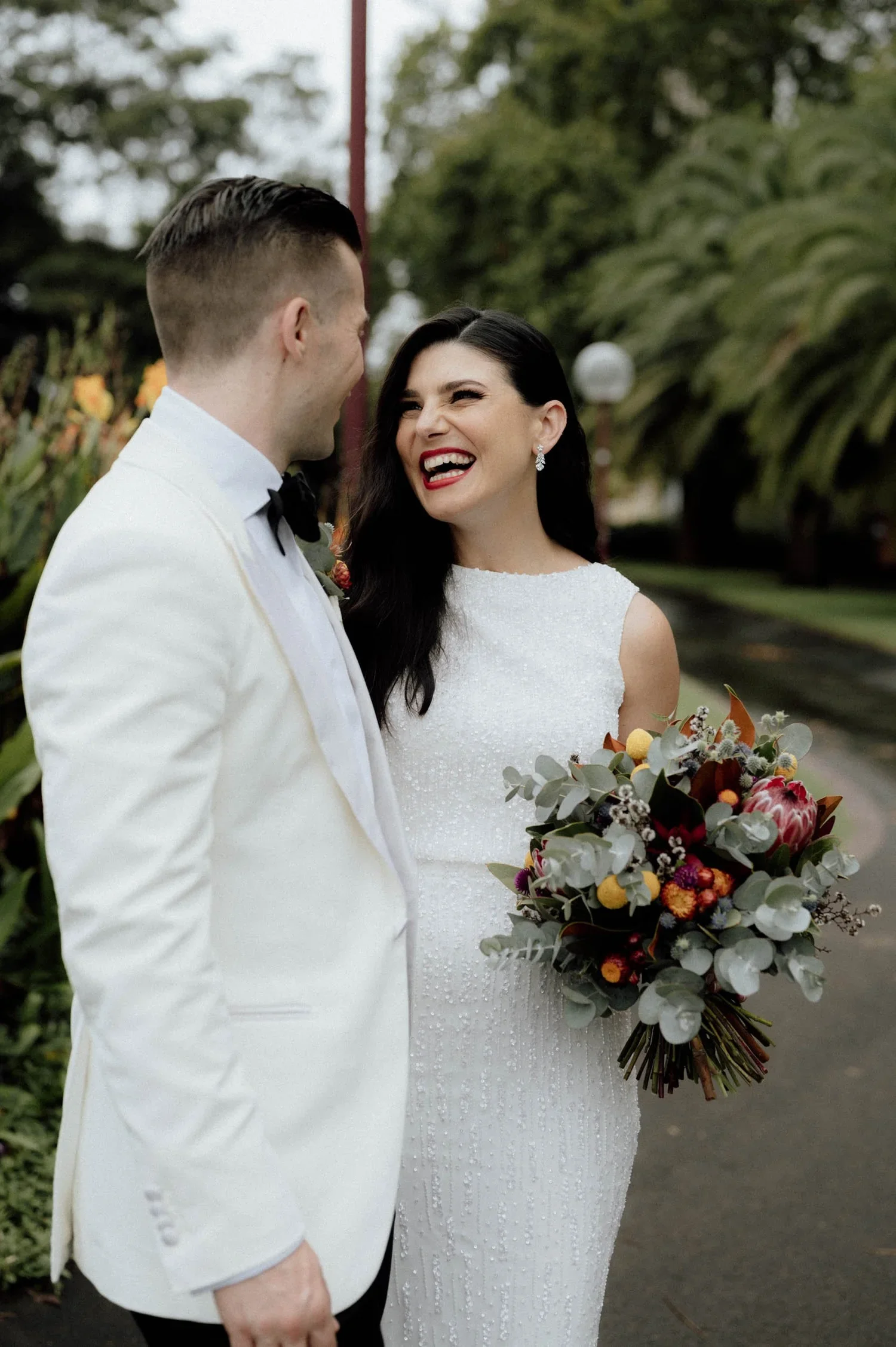 A bride and groom standing outdoors, smiling and looking at each other. The bride is holding a large bouquet of flowers with various colors and greenery. The groom is wearing a white tuxedo with a black bow tie. The background features trees and gree