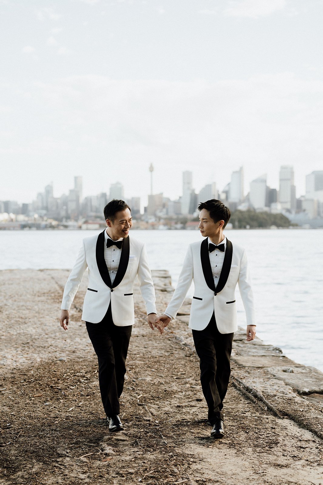 Two men in formal tuxedos holding hands and walking along a rocky waterfront with a city skyline in the background.