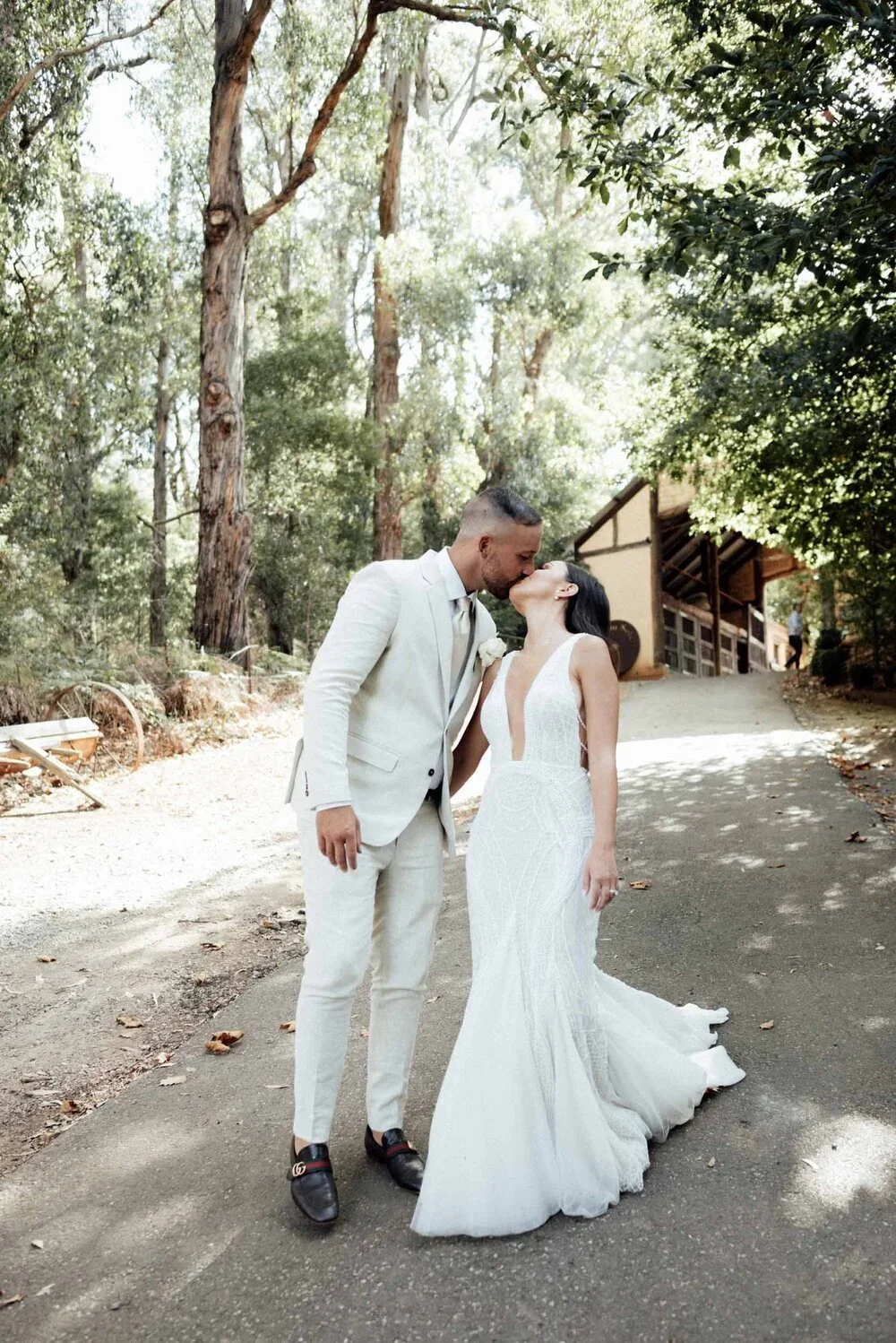 A couple dressed in wedding attire sharing a kiss outdoors among tall trees and greenery.