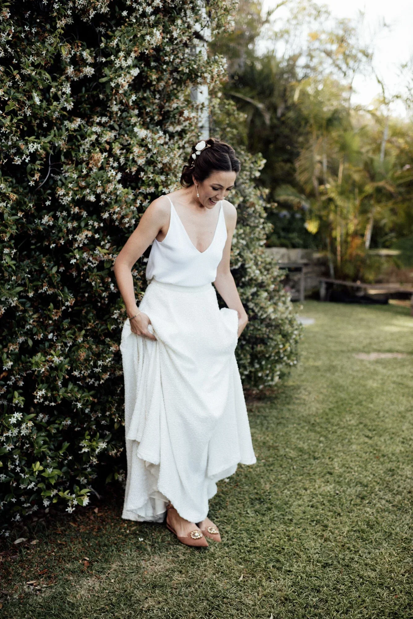 A woman in a white dress standing outdoors near a bush with small white flowers, smiling and slightly looking down, with greenery and trees in the background.