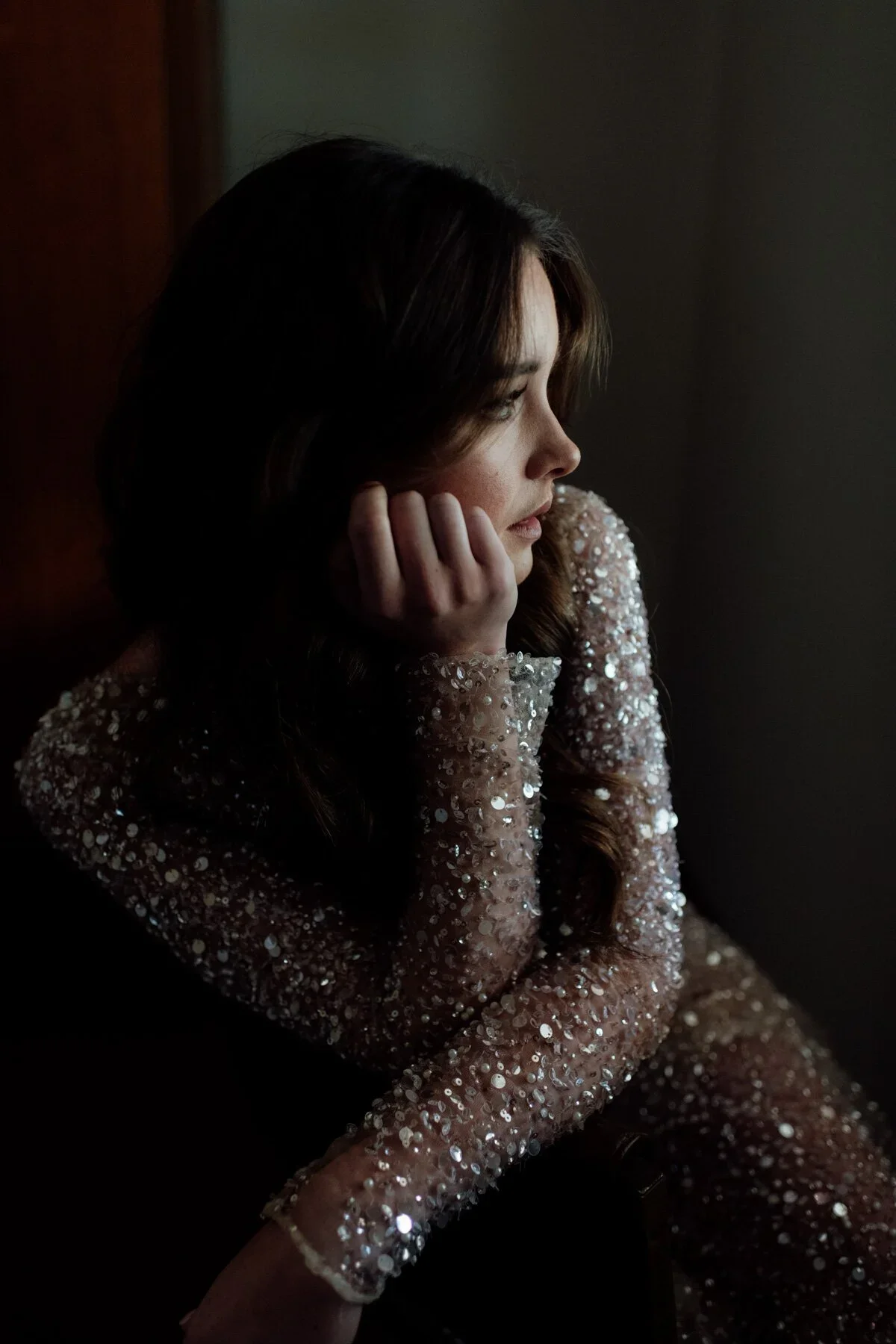 A woman with long dark brown hair looking to the right, resting her face on her hand, wearing a long-sleeved, beige dress covered in sequins, sitting in a dark room with soft light on her face.