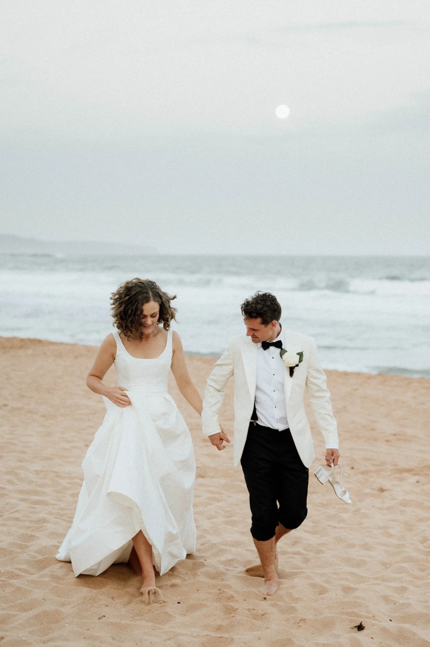 A newlywed couple walking barefoot on the beach holding hands during sunset, with the woman wearing a white wedding dress and the man in a tuxedo with a bow tie.