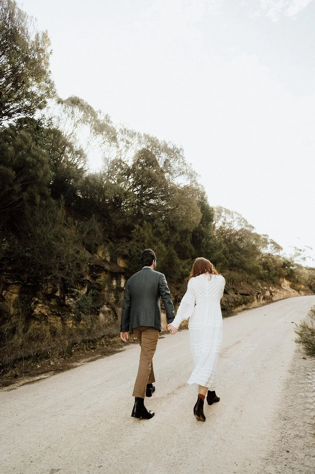A couple walking hand in hand on a dirt path surrounded by trees and bushes during daylight.