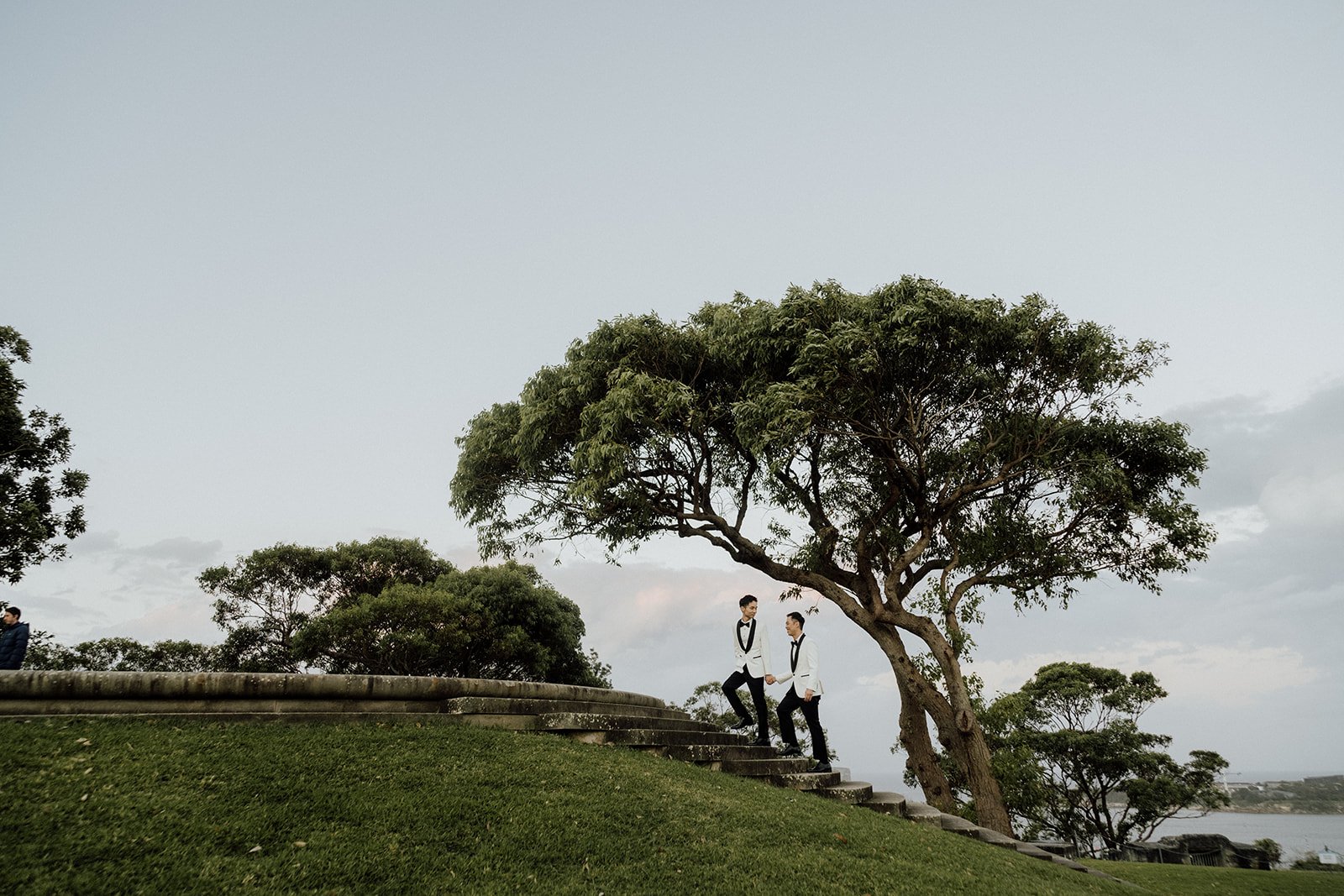Two people in tuxedos walking and holding hands on a curved set of stairs in a park with trees and a cloudy sky in the background, gunners barracks gay wedding.