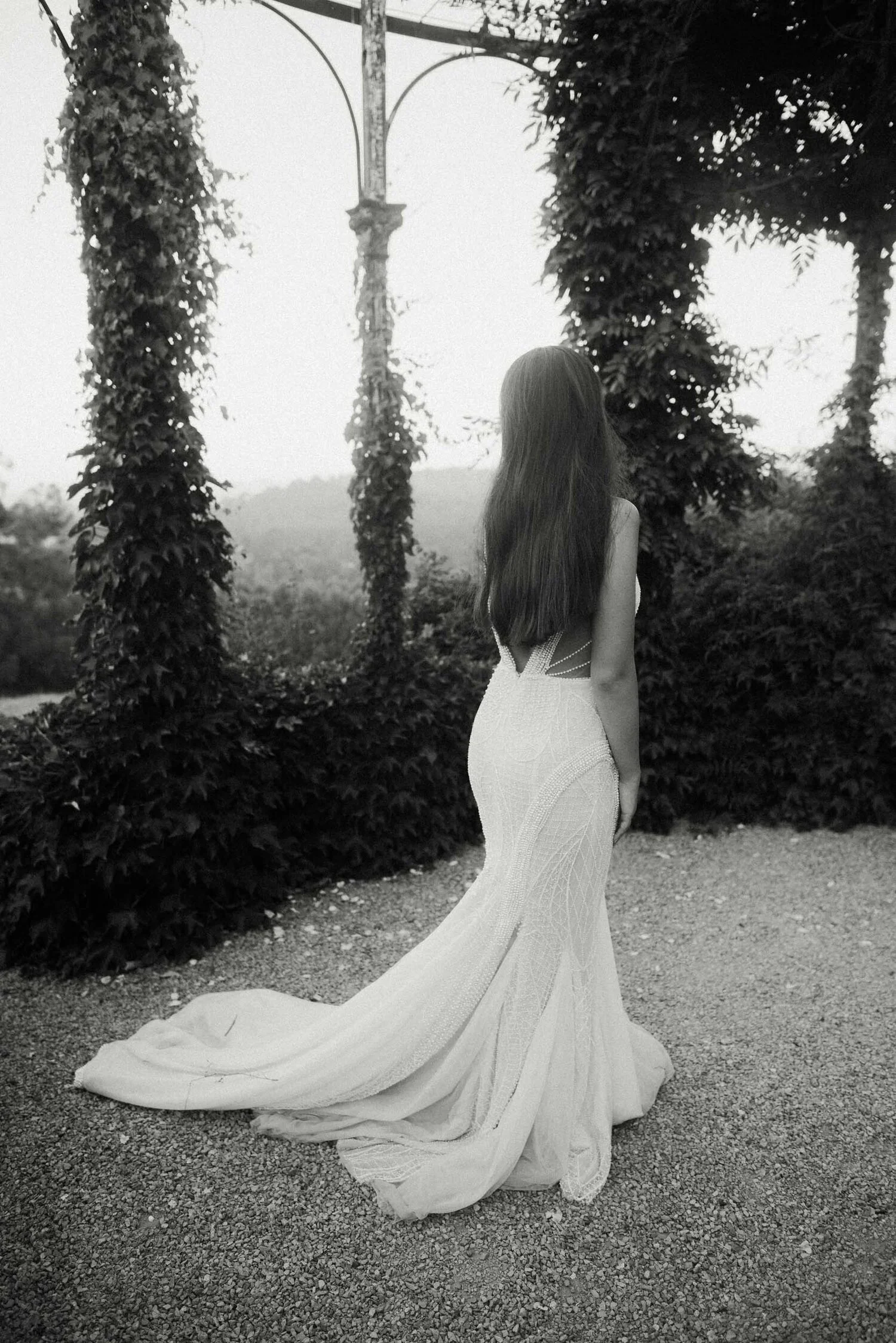 A woman in a strapless, form-fitting wedding dress with a long train, standing outdoors surrounded by greenery and foliage, facing away from the camera.