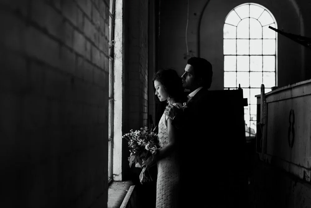 A black and white photo of a couple in wedding attire standing by a large window inside a church or historic building with high arched windows. The woman holds a bouquet of flowers, and the man gently embraces her from behind.