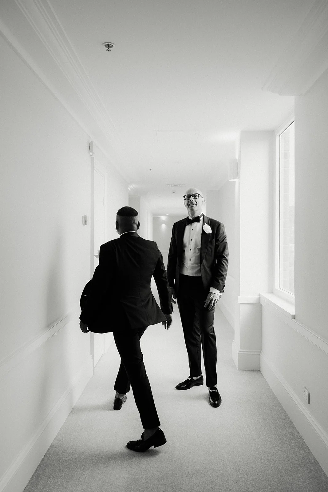 Two men in tuxedos walking down a bright, white hallway, smiling and talking to each other, they are grooms enjoying their same sex wedding in sydney.