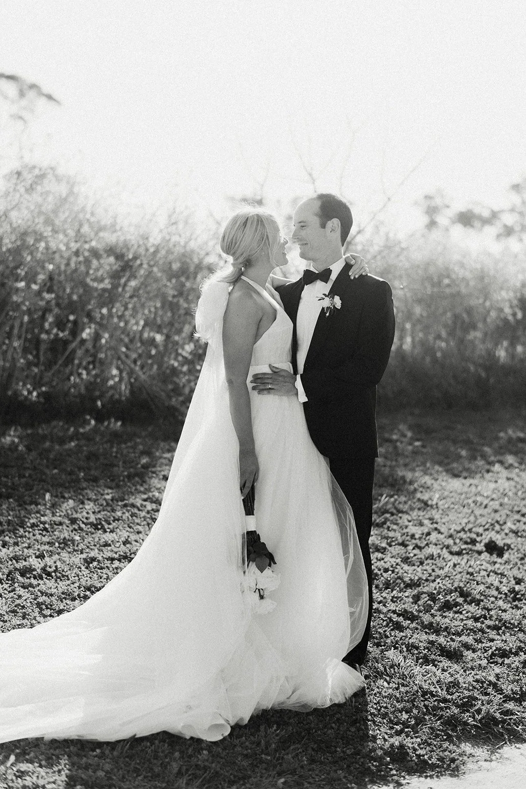 Black and white photo of a bride and groom smiling and looking at each other outdoors, with the bride holding a small bouquet of flowers.