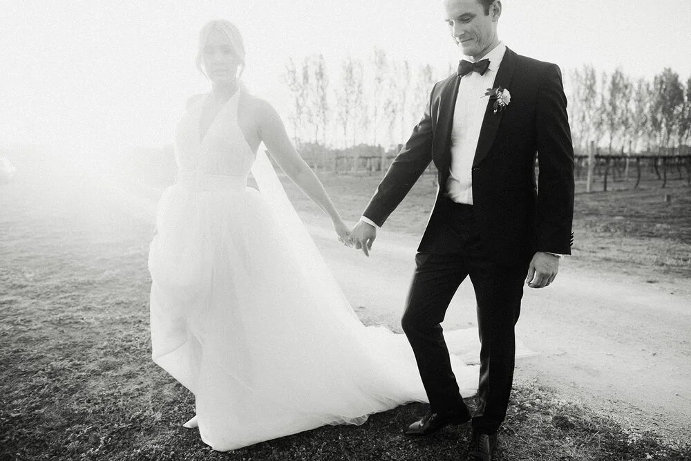 A black-and-white photo of a bride and groom holding hands outdoors, with the bride in a wedding gown and the groom in a tuxedo.