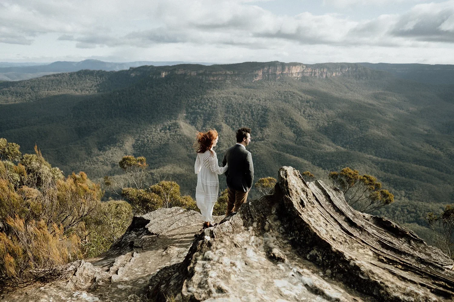 A man and woman standing on a rocky ledge overlooking a vast landscape of mountains and forests, with cloudy skies overhead.