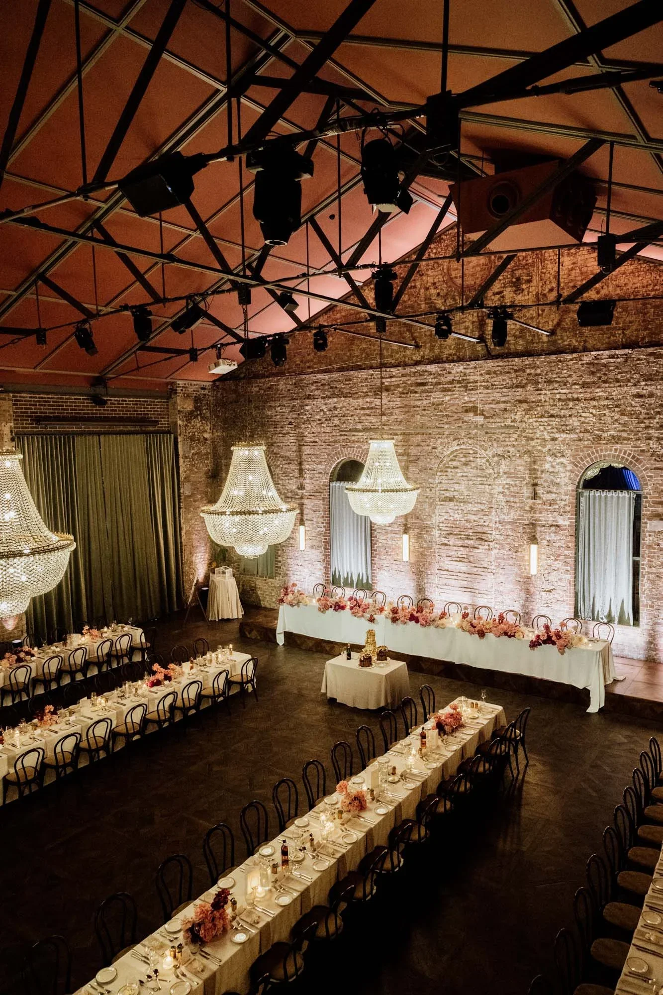 A large banquet hall decorated for an event, with long tables set for a meal, elegant chandeliers, and a stone brick wall backdrop.