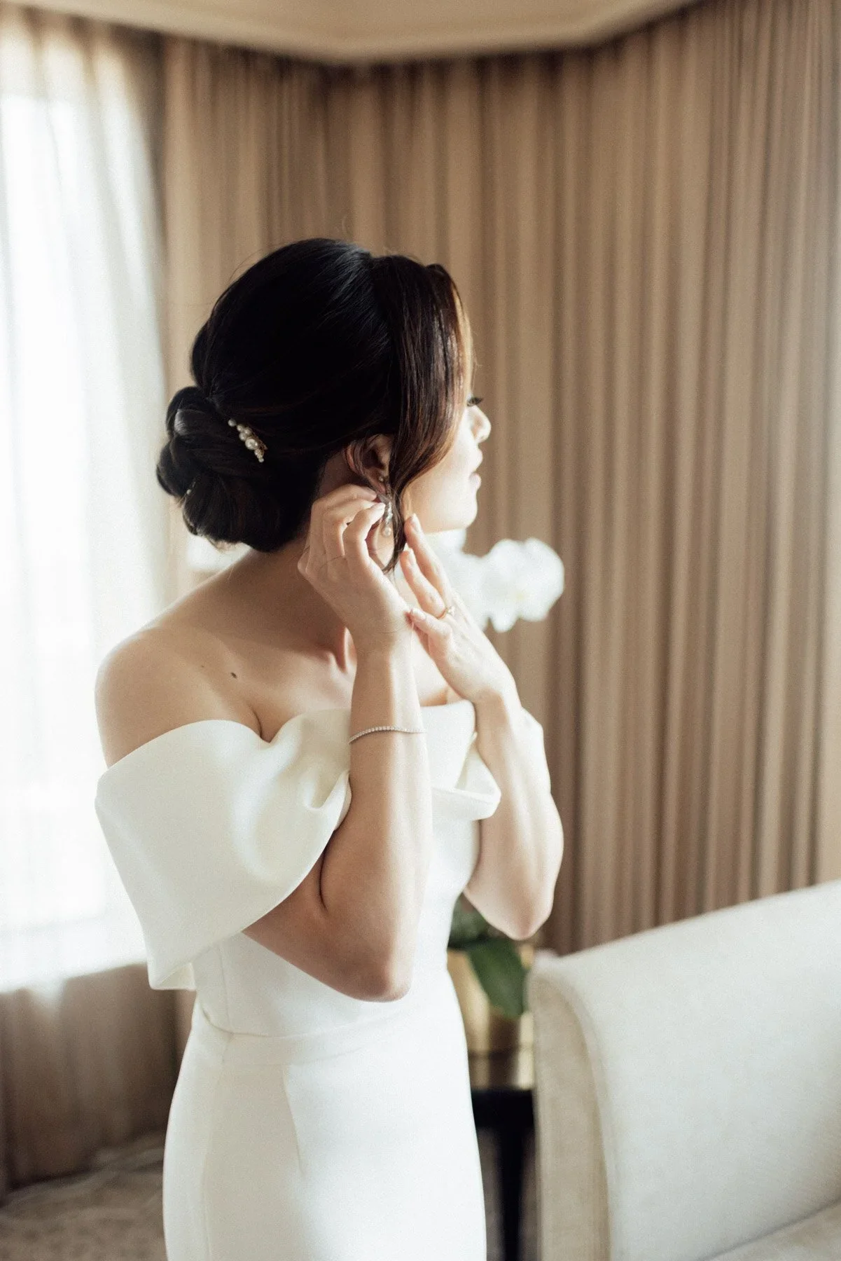 A woman in a white off-shoulder dress with puffed sleeves adjusting her earring in a room with beige curtains and a cream-colored chair.