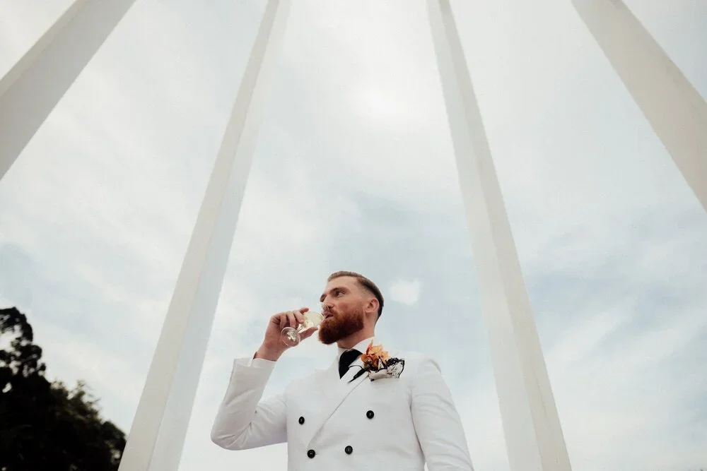 A man with a beard in formal white attire drinking champagne under large white arches.