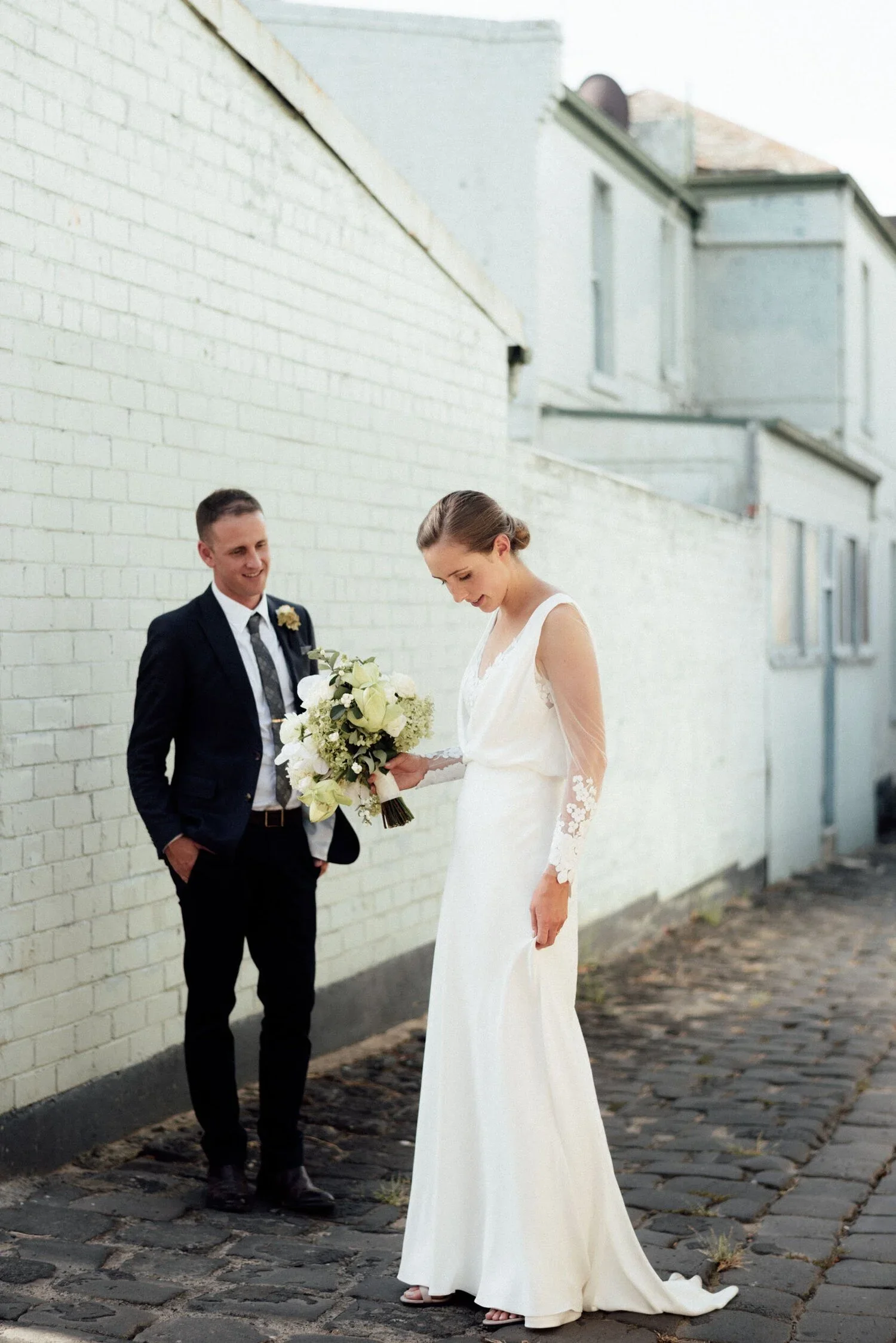 A bride in a white gown holding a bouquet of white flowers, looking down, standing on cobblestone street with a groom in a dark suit smiling at her, against a white brick wall.