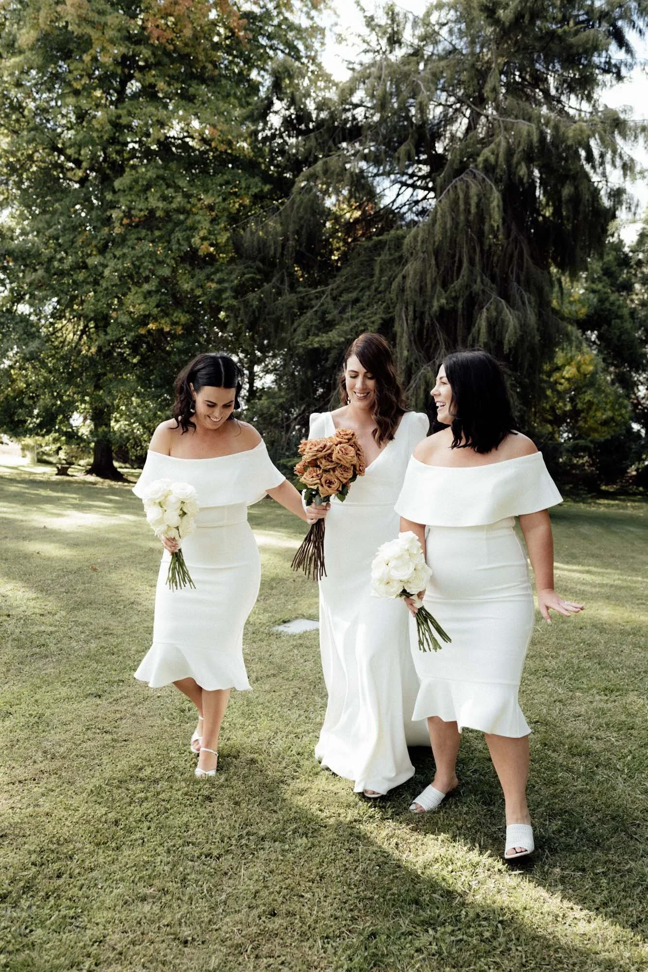 Three women in white dresses walking on grass in a park, carrying bouquets of flowers, smiling and enjoying themselves with trees in the background.