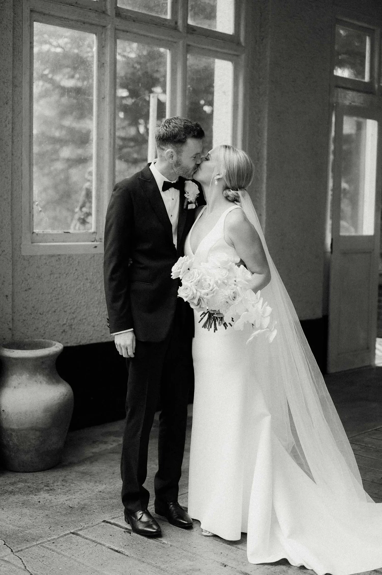 A newlywed couple is kissing indoors near a window. The groom is dressed in a tuxedo and the bride is wearing a white wedding gown and holding a bouquet of roses.
