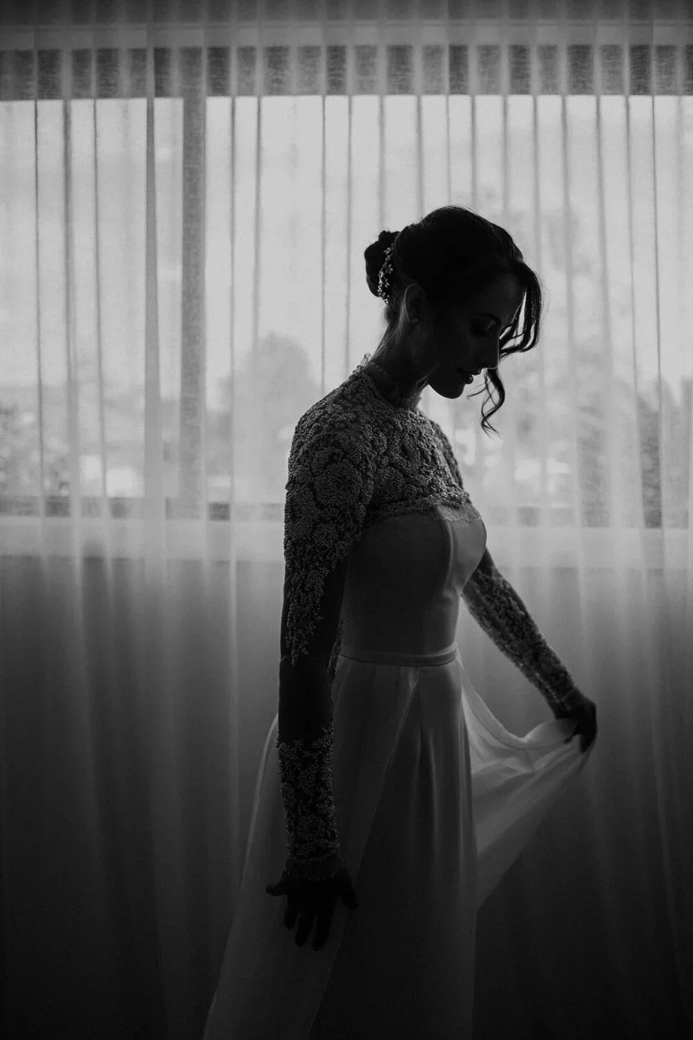 Black and white photo of a woman in a wedding dress standing near a window with sheer curtains, looking down and holding the fabric of her dress.